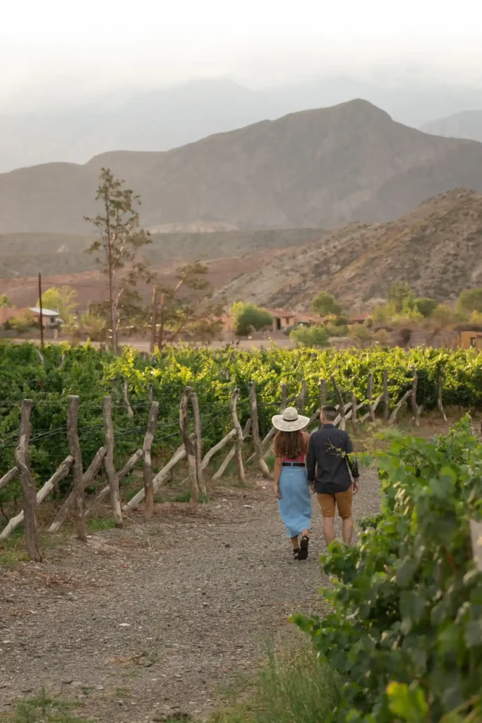 Casal caminha de mãos dadas em estrada de terra entre vinhedos e vinícolas de Mendoza, com a Cordilheira dos Andes ao fundo.